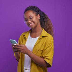 Young happy attractive African American woman using phone with smile to search for goods in internet store and online shopping at competitive prices stands in purple studio. Smartphone addiction